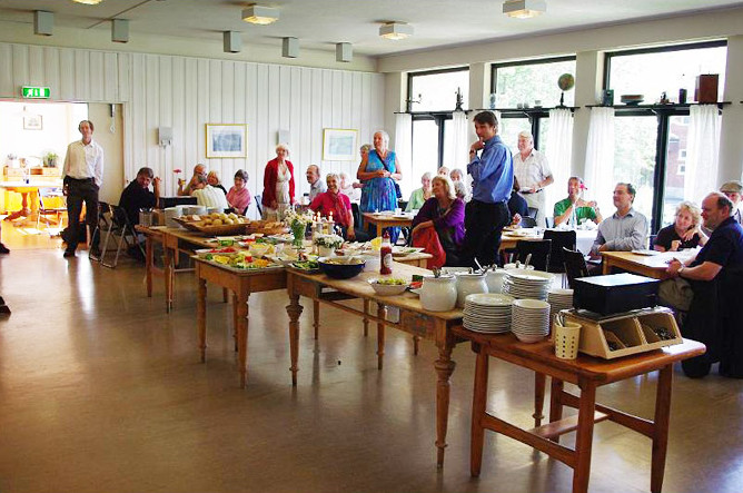 Lots of people are sat at tables, arranged around central tables on which there is a selection of food; everyone's attention is directed to a point behind and to the left of the camera