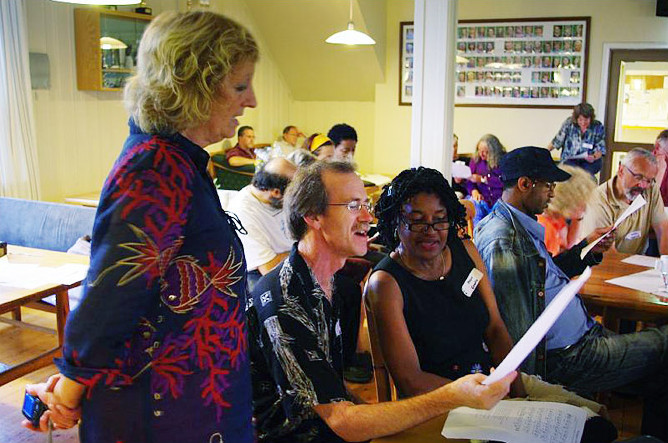 Three people, two seated and one standing, sing from a sheet